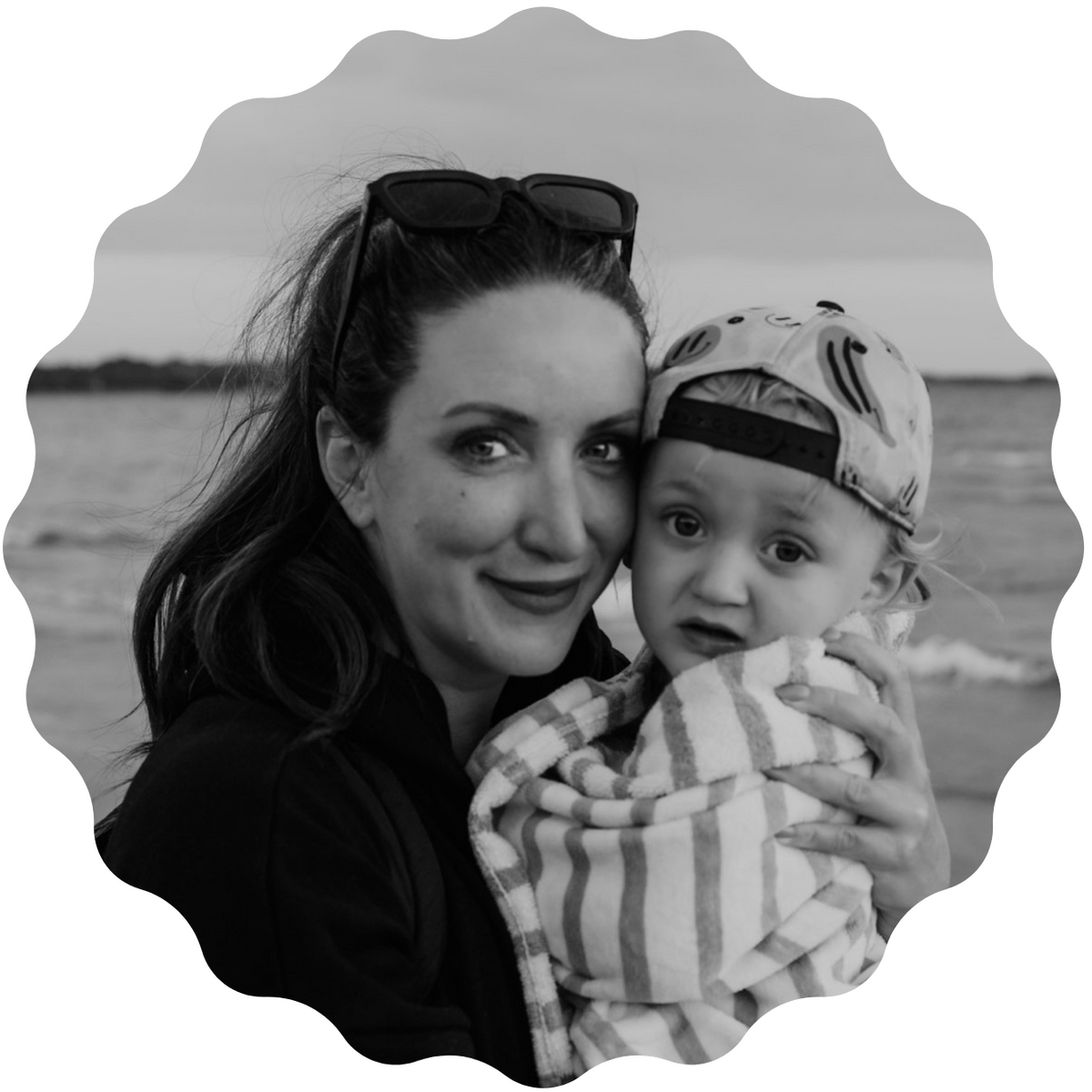 A black and white circular portrait of a woman holding a baby wrapped in a striped blanket, with sunglasses on her head and a blurred outdoor background.